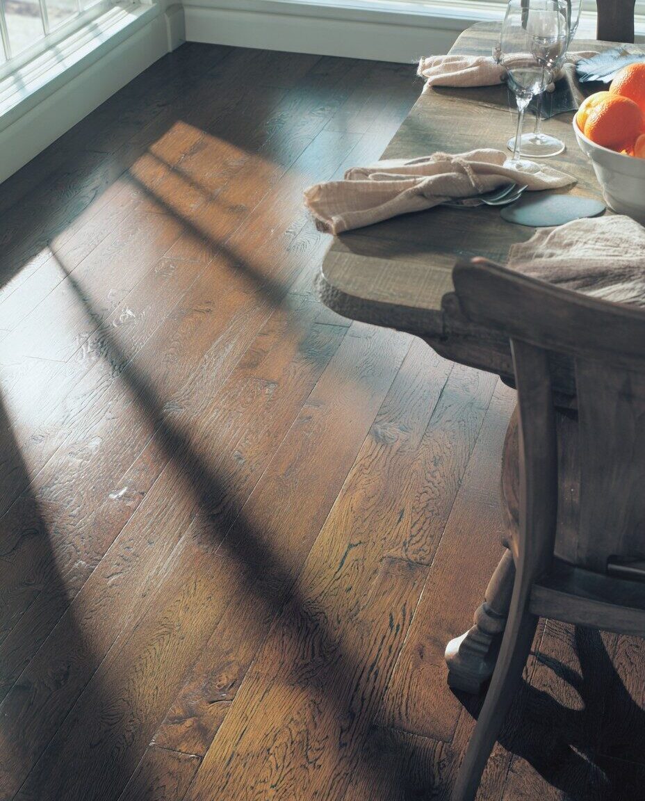 Dining room with rich brown hardwood flooring, wooden table set with napkins, glassware, and a bowl of oranges, with sunlight streaming through the window.